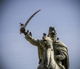 Dramatic low-angle view of a statue depicting a general and his horse with a raised sword against a clear blue sky backdrop, symbolizing strength and leadership.