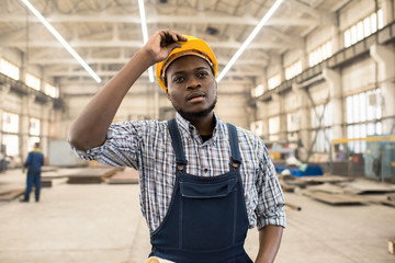 Portrait shot of young African American technician wearing overall and protective helmet standing at spacious production department of modern plant and controlling manufacturing process