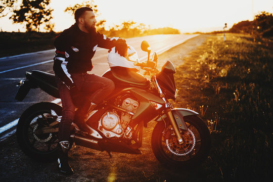 Full length portrait of an awesome young caucasian man sitting on his motorcycle while resting in his vacation trip near the road against sunset.