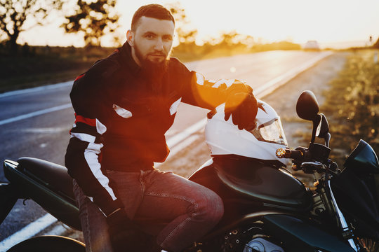 Portrait of a beautiful bearded man sitting near the road on his motorcycle while traveling around country looking into camera smiling against sunrise. © Strelciuc
