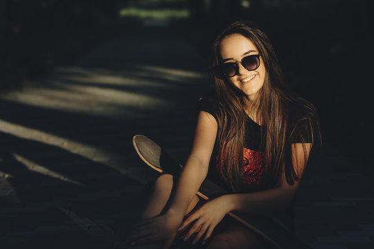 Portrait of a young long haired girl sitting in the park with a skateboard on her shoulders in direct sunlight with sunglasses and smiling at the photographer.