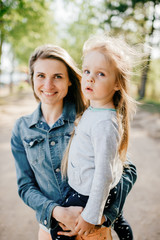 Happy smiling mother hugging her lovely little daughter outdoor. Lifestyle family. Adult cheerful female parent playing with her beautiful emotional child at nature in summer. Positive people faces.