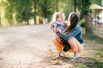 Happy smiling mother hugging her lovely little daughter outdoor. Lifestyle family. Adult cheerful female parent playing with her beautiful emotional child at nature in summer. Positive people faces.