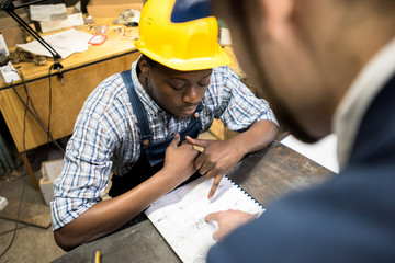 Young African American lathe operator wearing checked shirt and overall sitting at work bench and...