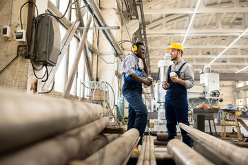 Multi-ethnic team of technicians wearing shirts and overalls gathered together at production department of modern plant with paper cups of coffee in hands and chatting with each other.