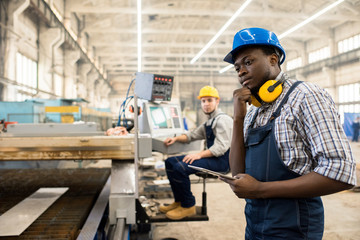 African American worker wearing overall and hardhat operating machine with help of digital tablet,...