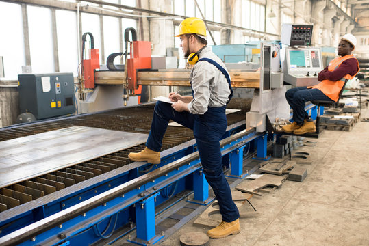 Confident inspector wearing hardhat and overall taking notes while carrying out inspection at production department of modern plant, African American technician sitting in front of CNC machine