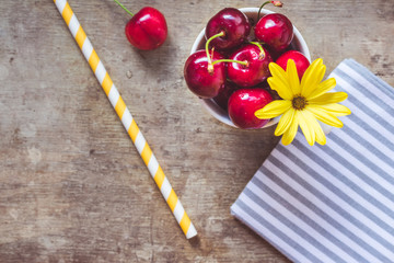 Close up and top view of fresh red cherries decorated with yellow flower in a white cup and a yellow straw on an rustic wooden background. Hello summer, weight loss or healthy diet concept