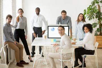 Young casual multiracial team and middle aged leader or coach looking at camera, smiling, standing near office desks in coworking space, posing for company business portrait. Team spirit, cooperation