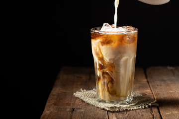 Ice coffee in a tall glass with cream poured over and coffee beans on a old rustic wooden table....