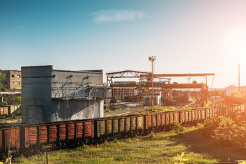 Fototapeta na wymiar Cargo freight train wagons go on railroad in industrial zone with plants and manufacturing factories of heavy industry at sunset light