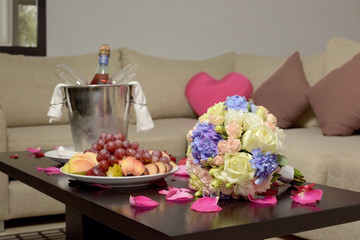 hotel room decorated for newlyweds, wooden table decorated with rose petals wedding bouquet fruits and a bucket for champagne in defocus