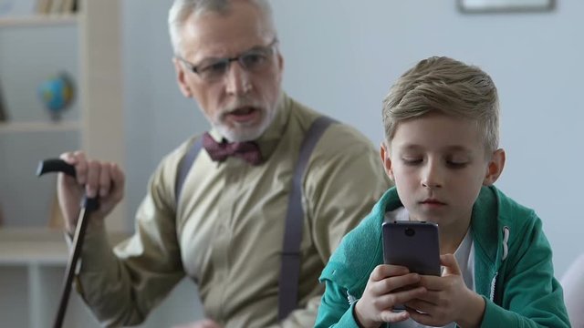 Angry old man scolding grandson playing smartphone game, family misunderstanding