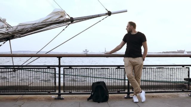 Young handsome man with backpack standing on the pier near the water close to bowsprit of the ship and looking around.