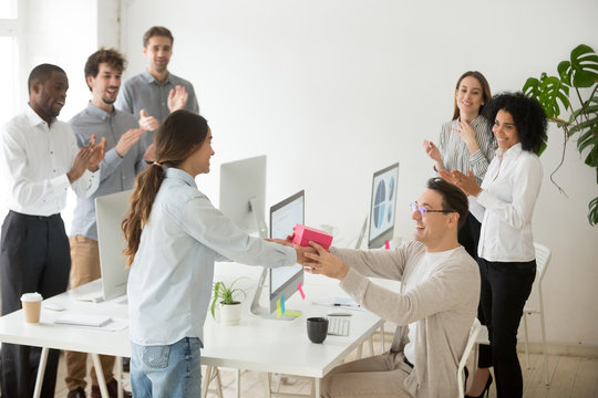 Smiling female employee congratulating colleague with birthday, presenting gift box, diverse coworkers greeting happy male manager, applauding and making unexpected surprise in office