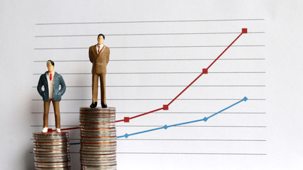 Miniature people standing on a pile of coins in front of a graph. The concept of the widening...