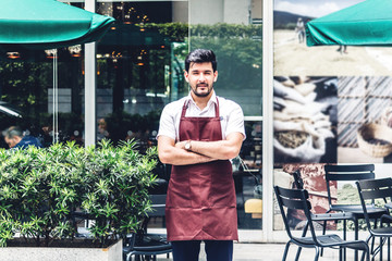 Portrait of handsome small business owner smiling and standing with crossed arms outside the cafe or coffee shop.Male barista standing at cafe