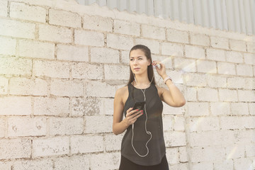 Athletic beautiful white female fitness model wearing long black sports wear with earphones connected to a mobile phone standing against a rough concrete  block wall background