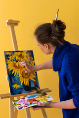 Girl artist paints sunflowers oil paints on canvas. She is wearing blue sweater. Woman is holding brush and palette with paints. She rubs paint with her finger.