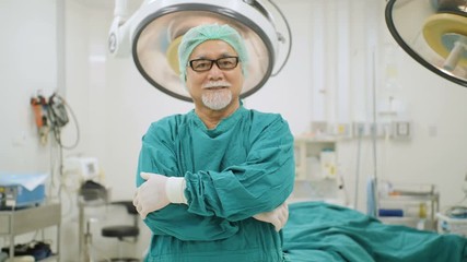 Portrait of senior male surgeon wearing full surgical scrubs smiling camera in operating theater at the hospital.