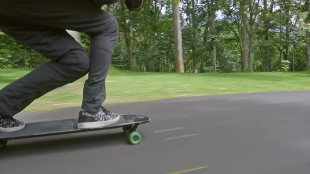 Follow low angle shot of young man in helmet leaning forward when skateboarding on road in green park on warm summer day