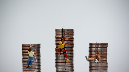 Miniature people climbing up a pile of coins with ropes....