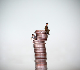 A miniature man and a miniature woman sitting on different heights from one stack of coins.