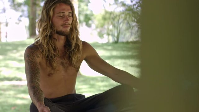 Young man with long and wavy fair hair sitting on bench in lotus pose, saying some words to himself and meditating with his eyes closed