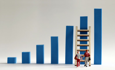 A miniature man and a miniature woman walking to a ladder with a blue bar graph. The...