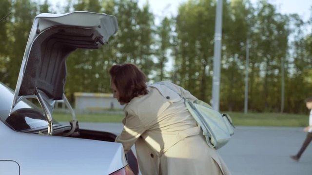 A woman in a beige raincoat opens the trunk of the car to get her suitcase.
