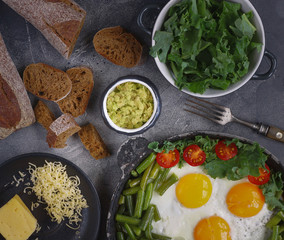 Fried eggs in an old pan with tomatoes, avocado pesto, green beans, pepper and salad on a dark background