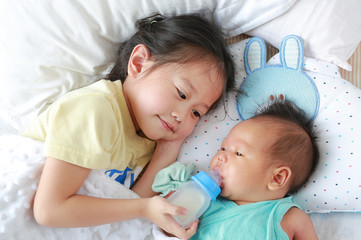 Cute Asian sister feeding milk from bottle for newborn baby lying on the bed.
