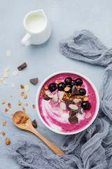 Pink black currant berries, banana smoothie bowl with granola, flakes from almonds and chocolate on light gray concrete background. Selective focus. Top view. Copy space.