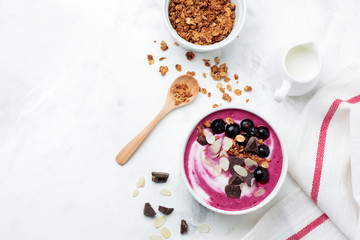 Pink black currant berries, banana smoothie bowl with granola, flakes from almonds and chocolate on light gray concrete background. Selective focus. Top view. Copy space.