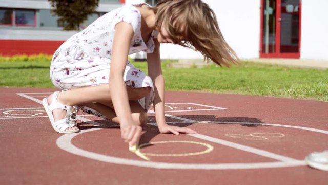 Little cute girls draws Hopscotch on asphalt by chalk. Children playing on playground outdoors. Kids jumps hopscotch at park. 