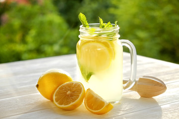 Fototapeta na wymiar One mason jar glass of homemade lemonade, slices of organic ripe lemon, whole and halved, mint, juicer on white wooden table, country side tree foliage background. Close up, top view, copy space.