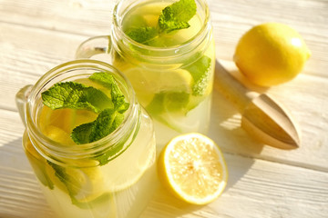 Two mason jar glasses of homemade refreshing lemonade, slices of organic ripe lemon, whole and halved, mint leaves, juicer, muddler, squeezer on a rustic white wooden background. Close up, top view.