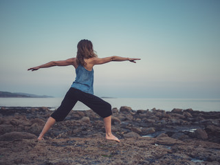 Fototapeta premium Woman in yoga warrior pose on coast at sunset