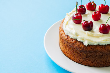 Homemade sour cream cake with cream cheese frosting decorated with fresh ripe cherries on a light blue background. Close up and copy space.