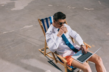 high angle view of young businessman in shorts working with laptop while sitting on sun lounger on asphalt and drinking coffee to go