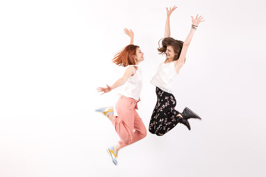 Two Happy And Positive Women Jump Over A White Wall In Studio