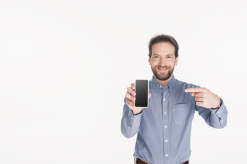 portrait of smiling bearded man pointing at smartphone with blank screen in hand isolated on white