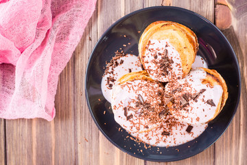 A pile of pancakes stuffed with fruit yoghurt and sprinkled with grated chocolate on a plate on a wooden table. Top view