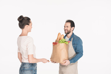 side view of woman giving credit card to shop assistant in apron with paper package full of food isolated on white