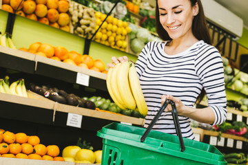 Young woman chooses bananas at the store and put them in a basket for the products.