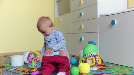 Smiling baby sitting on chamber pot with lots of toys and toilet paper around him in kids room