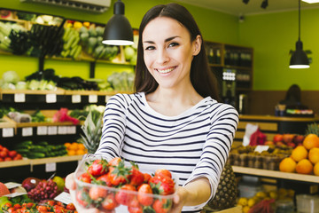 Beautiful young smiling woman holding a box with fresh strawberries and showing at the camera in a supermarket