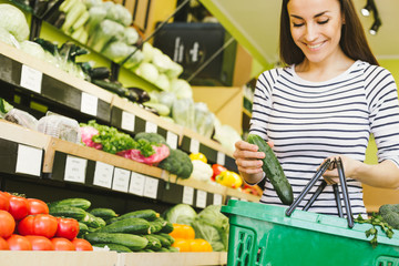 The choice of vegetables in the supermarket. Smiling beautiful woman puts a cucumber in a food basket on a background of vegetables and fruits