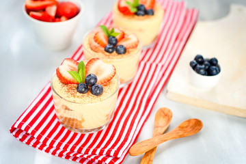 homemade, exquisite dessert tiramisu in glasses decorated with strawberry, blueberry, mint on white wooden table, top view.