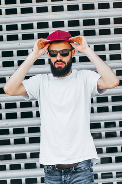 A stylish man with a beard in a white T-shirt and glasses and cap on a wall background. Street photo